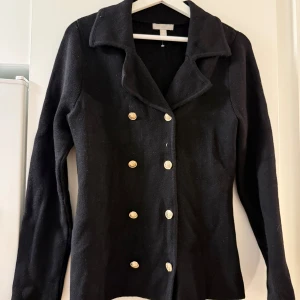 Svart cardigan jacka från H&M, S - Svart stickad cardigan jacka från H&M i storlek S. Jackan har dubbelknäppning med guldiga knappar framtill och en klassisk krage. Långärmad och figurnära modell, perfekt för en stilren look.