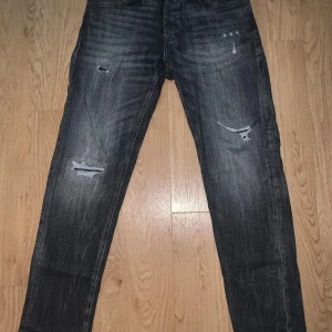 Svarta ripped jeans Jack & Jones - Säljer ett par svarta jeans från Jack & Jones i modellen Tapered/Mike. Jeansen har snygga slitningar och en tvättad look som ger dem en cool vibe. Passformen är tapered med smalare ben och de är tillverkade i klassiskt jeansmaterial. Perfekta för dig som gillar streetstyle.