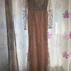 Lång vit mönstrad klänning - Säljer en långklänning i vitt med transparenta långärmar och geometriskt mönster. Klänningen har en figurnära passform och är dekorerad med glittriga detaljer som ger en lyxig känsla. Perfekt för dig som vill sticka ut med en unik look.