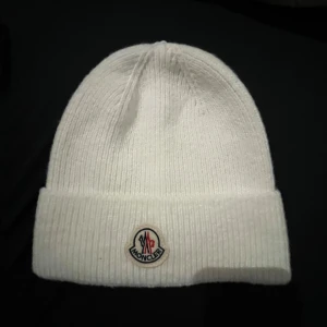 Vit stickad mössa från Moncler - Säljer en stilren vit stickad mössa från Moncler med klassisk uppvikt kant och broderad logotyp framtill. Mössan är ribbstickad och passar perfekt för kyliga dagar. Enkel och clean design som funkar till det mesta.