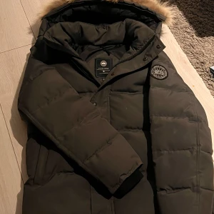 Mörk grå Canada Goose jacka, Black lable - Säljer en mörk grå dunjacka från Canada Goose med klassisk patch på ärmen och stor huva med päls. Jackan är tjock, har dragkedja och knappar framtill samt två fickor. Perfekt för kalla vinterdagar och riktigt snygg streetstil. Avtagbar päls
