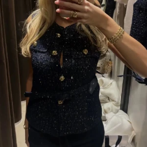 Mörkblå glittrig blus med bälte - Säljer en mörkblå glittrig blus i tweed-liknande material med guldfärgade knappar och tillhörande bälte i midjan. Blusen är ärmlös och har två fickor framtill med dekorativa knappar. Perfekt för dig som vill ha en chic och lyxig vibe.