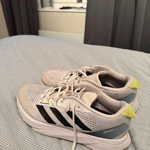 Adidas Adizero SL vita sneakers - Snygga Adidas Adizero SL sneakers i vitt med svarta ränder och detaljer i ljusgul och grå. Skorna har ventilerande meshmaterial, snörning och platt sula. Perfekta för dig som gillar sportig stil och vill ha bekväma skor till träning eller vardag.