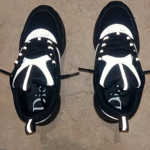 Svarta Dior sneakers med silverdetaljer - Säljer ett par chunky Dior sneakers i svart mesh och skinn med silvriga detaljer och reflekterande partier. Skorna har snörning, platt sula och rund tå. Märkeslogga på sidan och innersulan. Perfekt för dig som gillar statement-sneakers med lyxig vibe.