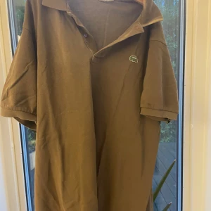 Brun pikéskjorta från Lacoste - Säljer en brun pikéskjorta från Lacoste med klassisk krage och korta ärmar. Skjortan har den ikoniska gröna krokodilloggan broderad på bröstet och knappar vid halsen. Materialet är mjuk bomull som känns skönt mot huden.