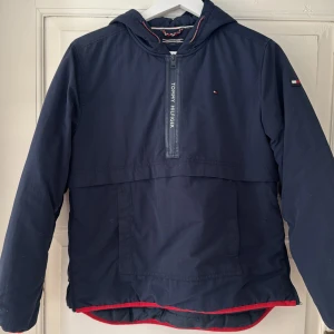 Snygg Mörkblå vinterjacka Tommy Hilfiger 164 - Snygg mörkblå vinterjacka från Tommy Hilfiger med huva, halv dragkedja framtill och logga på bröstet. Jackan har röda och vita detaljer på huva och nederkant samt praktiska fickor. Perfekt för dig som gillar sportig och stilren look.