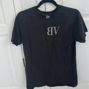 Svart t-shirt med strass från Bvaldi - Svart t-shirt från Bvaldi i bomull med korta ärmar. Framsidan har ett glittrigt VB-tryck i strass och baksidan har BVALDI i stora strassbokstäver. Klassisk passform och rund halsringning. Perfekt för dig som gillar statement-plagg. Storlek S skicka 8/10 inga defekter 