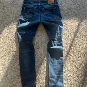 Blå slim fit jeans från Nudie Jeans - Säljer ett par blå skinny jeans från Nudie Jeans med klassisk femficksdesign, orangea sömmar och Nudie-logga på bakfickan. Jeansen har en mörk tvätt med snygga slitningar och är tillverkade i stretchig denim för en tight passform.