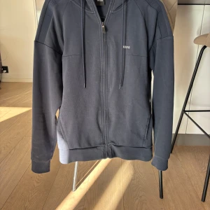 Mörkblå hoodie från BOSS med dragkedja - Snygg mörkblå hoodie från BOSS med hel dragkedja framtill, huva med snörning. Väldigt fint skick. Hör av er om ni har några funderingar.