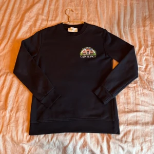 Svart sweatshirt från Casablanca - Svart sweatshirt från Casablanca med färgglad broderad grafik och logga på bröstet samt stor illustration på ryggen. Tröjan har rund hals, långa ärmar och mjukt material som känns skönt mot huden. Perfekt för dig som gillar unika prints och streetstyle.