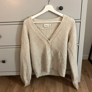 Beige ribbad kofta från Kappahl - Mysig beige kofta från Kappahl i storlek S. Koftan är ribbstickad med två knappar framtill, v-ringad och har långa ärmar. Perfekt för lager-på-lager och chill dagar. Enkel att matcha med jeans eller kjol.