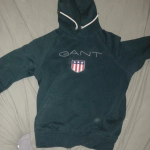 Mörkgrön hoodie från GANT med brodyr - Snygg mörkgrön hoodie från GANT med broderad logga och sköld på bröstet. Tröjan har huva med vita snören och en stor magficka. Perfekt för dig som gillar klassisk och sportig stil.