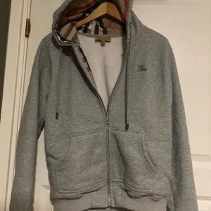 Burberry zip-up hoodie  - Grå hoodie från Burberry i bomull med klassiskt rutigt foder i huvan. Tröjan har dragkedja framtill, två fickor och broderad logga på bröstet. Perfekt för dig som gillar stilren design med en touch av lyx.