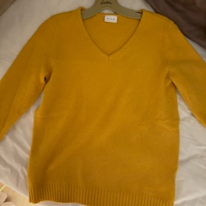 Gul stickad tröja från Vila - Mysig gul stickad tröja från Vila med v-ringad hals och ribbade muddar. Perfekt för lager-på-lager och ger en färgklick till din outfit. Långärmad och i mjukt material som känns skönt mot huden.