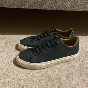 Mörkblå sneakers från Zara - Säljer ett par stilrena mörkblå sneakers från Zara med vit sula och blåa snören. Skorna har en klassisk låg profil och är fodrade med beige insida. Perfekta för dig som gillar enkel och clean stil strl 39-39,5