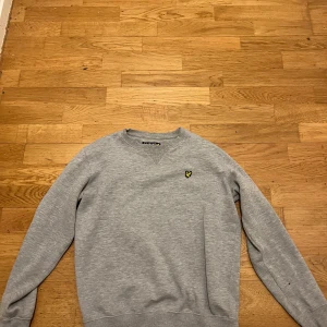 Grå sweatshirt från Lyle & Scott - Snygg grå sweatshirt från Lyle & Scott med klassisk rund hals och broderad gul logga på bröstet. Tröjan har ribbade muddar vid ärmslut och midja, perfekt för en chill och stilren look.
