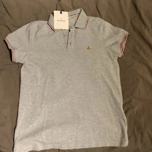 Grå Moncler pikétröja med logga - Snygg grå pikétröja från Moncler med klassisk krage och knäppning framtill. Tröjan har korta ärmar med röd och marinblå rand på ärmslut och krage samt broderad Moncler-logga på bröstet. Tillverkad i mjuk bomull för en skön känsla.