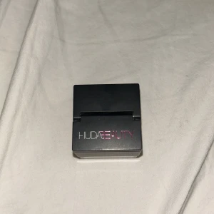 Huda Beauty CHERRY BLOSSOM CAKE med puff - Säljer ett superfint glittrigt puder från Huda Beauty i en smidig svart och transparent förpackning. Kommer med en mjuk puff för enkel applicering och har en söt ljusrosa som lyser upp under ögonen. Perfekt att ha med i väskan för touch-ups. Köpt inne på Sephora!