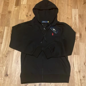 Svart hoodie från Polo Ralph Lauren - Svart hoodie från Polo Ralph Lauren med dragkedja och huva. Klassisk röd broderad logga på bröstet och snörning i huvan. Tillverkad i mjuk bomullsmix, perfekt för chill dagar. Snygg och enkel att matcha med allt.