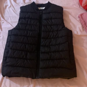 Svart dunväst från H&M - Svart dunväst från H&M i storlek 146/152. Västen har quiltad design med horisontella sömmar och dragkedja framtill. Perfekt att slänga över en hoodie för extra värme. Enkel och stilren look som funkar till det mesta.