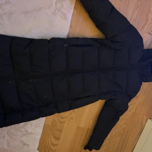 Svart lång pufferjacka från Áhkká - Svart lång pufferjacka från Áhkká Outdoor med huva och diskret logga på ärmen. Jackan har quiltad design, dragkedja framtill och är perfekt för kalla dagar. Materialet är syntet och jackan har långa ärmar. Snygg och funktionell för vinter och höst.