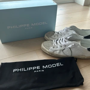 Philippe Model vita sneakers med mockadetaljer - Snygga vita Philippe Model sneakers med klassisk låg profil och rund tå. Skorna har ovandel i skinn med mockadetaljer på tungan och framdelen, samt svarta detaljer på hälen. Platt sula och snörning framtill. Perfekt för dig som gillar stilrena och trendiga sneakers. Storlek 40 mvh Filip.