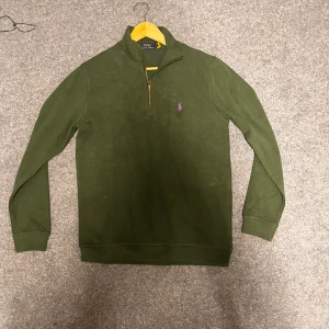 Grön half zip Polo Ralph Lauren - Snygg mörkgrön långärmad tröja från Polo Ralph Lauren med lila broderad logga på bröstet. Tröjan har en half zip-dragkedja och är tillverkad i mjuk bomull. Perfekt för lager-på-lager och chill stil.