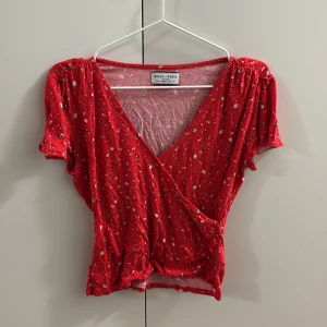 Röd blommig omlottopp Holly & White - Säljer en röd omlottopp från Holly & White med blommigt mönster. Toppen har korta ärmar, v-ringning och är croppad med lätt rynk vid axlar och midja. Perfekt för dig som gillar färg och mönster!