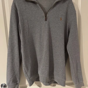 Grå half zip tröja från Polo Ralph Lauren - Snygg grå långärmad tröja från Polo Ralph Lauren med half zip och klassisk broderad logga på bröstet. Tröjan har en normal passform och är tillverkad i mjuk bomull, perfekt för lager-på-lager under höst och vinter.