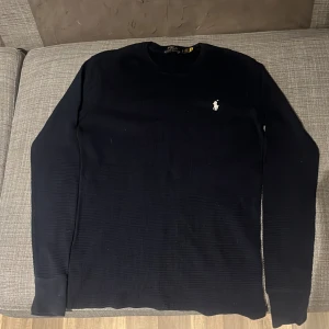 Svart långärmad tröja Polo Ralph Lauren - Svart långärmad tröja från Polo Ralph Lauren med klassisk rund hals och vit broderad logga på bröstet. Tröjan är ribbad och tillverkad i mjuk bomull, perfekt för lager-på-lager under kyliga dagar. Använd nån enstaka gång
