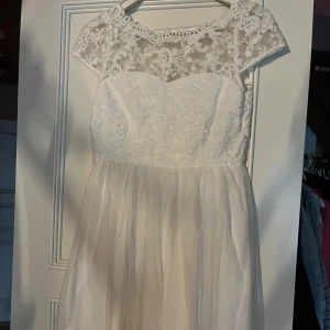 Vit spetsklänning med öppen rygg - Superfin vit kortklänning med spets upptill och transparenta detaljer vid axlar och rygg. Kjolen är lätt och faller snyggt med ett tunt lager tyg. Klänningen har korta ärmar och v-ringad öppen rygg, perfekt för student eller sommar. Använd 1 gång.