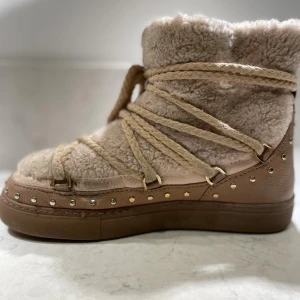 Beige inukkis med fårull och nitar - Säljer ett par beige innukis med mjuk fårull och grova snören som går runt hela skon. Skorna har bruna detaljer i skinn och är dekorerade med små silver- och guldfärgade nitar längs sulan. Perfekta för kalla dagar och riktigt mysiga att ha på sig. Endast använda några gånger förra vintern 