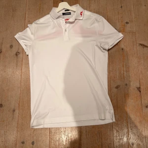 Vit J.Lindeberg piké med röd detalj - Vit pikétröja från J.Lindeberg med klassisk krage och knappar framtill. Snygg röd logodetalj på kragen och en stor röd grafisk detalj över ryggen. Tillverkad i mjukt och stretchigt syntetmaterial, perfekt för en sportig look.