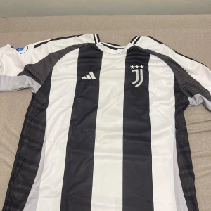 Juventus Vlahovic #9 fotbollströja M - Juventus matchtröja från Adidas med Vlahovic och nummer 9 på ryggen. Klassisk svartvit randig design, Serie A-märke på ärmen och klubbens emblem med tre stjärnor på bröstet. Tillverkad i lätt polyester, perfekt för fotboll eller att representera ditt lag.