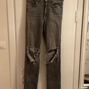 Gråa jeans från Zara - Ett par mörkgråa jeans från Zara i storlek 40. För referens så är jag 172 cm och jeansen går ner till marken för mig. Det är en tajt modell med mellan midja som har slits vid fotändan på båda benen, vilket ger en stilren look där jeansen faller över skorna snyggt. Det är hål vid knäna, hålen är relativt stora och faller snyggt vid knäna. Det finns några få slitningar i form av trådar (dessa går att klippa bort bara)