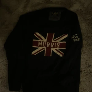 Svart stickad tröja Morris Union Jack - Svart stickad tröja från Morris med Union Jack-flagga och texten 'Morris' på bröstet. Rund halsringning och lång ärm. Snygg detalj med broderad text på ena ärmen. Perfekt för dig som gillar klassisk brittisk stil.