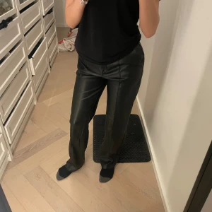 Svarta raka byxor i skinnimitation NA-KD - Snygga svarta byxor från NA-KD i skinnimitation med raka ben och normal passform. Byxorna har klassiska fickor bak och stängs med dragkedja och knapp framtill. Perfekta för en trendig look och lätta att matcha med olika toppar.