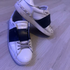 Valentino Garavani Open sneakers blå/svart - Säljer ett par Valentino Garavani Open sneakers i vitt skinn med bred svart läderdetalj över vristen. Klassisk rund tå, snörning och platt sula. Ikoniska nitar baktill på sulan ger extra edge. Perfekta för dig som gillar lyxiga och stilrena sneakers.