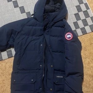 Mörkblå dunjacka från Canada Goose - Säljer en mörkblå dunjacka från Canada Goose med huva och klassisk logga på ärmen. Jackan har stora fickor med knappar, dragkedja och tryckknappar framtill. Perfekt för kalla vinterdagar och riktigt snygg streetstil.