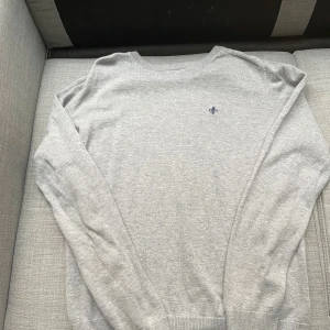 Ljusgrå stickad tröja från Morris - Säljer en stilren ljusgrå stickad tröja från Morris i 100% pima bomull. Tröjan har rund halsringning, ribbade muddar och en liten broderad logga på bröstet. Perfekt för dig som gillar klassisk och clean stil.