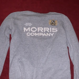 Grå stickad tröja från Morris Company - Snygg grå stickad tröja från Morris Company med broderad logga och text framtill. Tröjan har rund halsringning och långa ärmar, perfekt för lager-på-lager-stil. Klassisk och stilren design som funkar till det mesta. Hyfsat stor i storlek