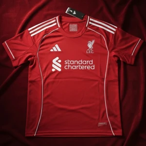 Liverpool  fotbollströja  - Röd Liverpool fotbollströja från Adidas med vita detaljer och klubbmärke på bröstet. Klassiska tre ränder på axlarna och Standard Chartered-logga framtill. Tillverkad i lätt och ventilerande material, perfekt för match eller träning.