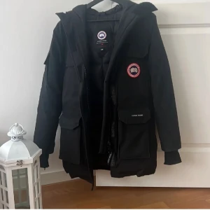 Svart dunjacka Canada Goose XS - Svart dunjacka från Canada Goose med klassisk patch på bröstet och stora fickor framtill. Jackan har huva, dragkedja och är tillverkad av polyester och bomull med dunfyllning. Perfekt för kalla vinterdagar och har en snygg, stilren look.