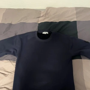 Mörkblå sweatshirt från Stone Island - Säljer en mörkblå sweatshirt från Stone Island med klassisk rund hals och ikonisk logga på vänster ärm. Tröjan har en clean look och är tillverkad i mjuk bomull, perfekt för dig som gillar streetwear och stilren design. Storlek M men passar även S, tröjan är i ett jättebra skick, knappt använd och inga defekter eller sådant!