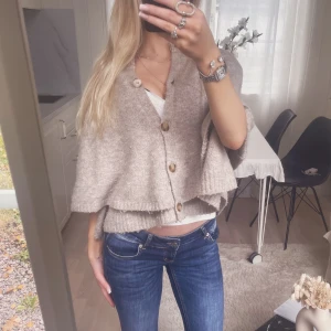 Beige stickad kofta med knappar - Supermysig höstig beige stickad kofta med stora träknappar och lös passform. Koftan är som en poncho, perfekt att slänga över ett linne eller t-shirt. Enkel att styla till både jeans och kjol. Den är lite nopprig