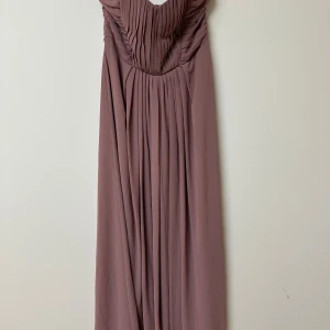 Lång plommonfärgad klänning H&M - Elegant långklänning från H&M i en mjuk plommonlila nyans. Klänningen är axelbandslös med vackra draperade detaljer upptill och har en lätt utställd kjol. Tillverkad i ett tunt, följsamt material och stängs med dragkedja bak.