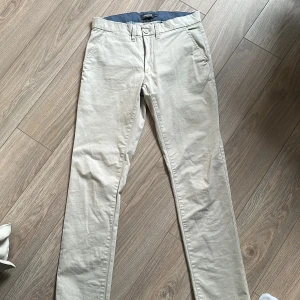 Beige chinos från Dressmann slim fit i storlek 31/32 - Snygga beige chinos från Dressmann i slim fit-modell. Byxorna har klassisk design med framfickor, bakfickor med knapp och bälteshällor. Tillverkade i mjukt bomullsmaterial som är skönt mot huden. Perfekt för dig som gillar stilrena och enkla byxor.
