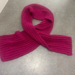 Mörkrosa stickad halsduk - En chunky, ribbstickad halsduk i mörkrosa färg. Halsduken är ca 18 cm bred och 150 cm lång, perfekt att vira flera varv runt halsen. Den grova stickningen ger en cool och mysig vibe, och färgen poppar verkligen till vilken outfit som helst.