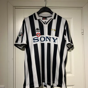 ÄKTA Juventus Zidane 21 Kappa fotbollströja - Säljer en äkta Juventus fotbollströja med klassiska svarta och vita ränder, Kappa-logga, Sony som sponsor och Lega Calcio-märke på ärmen. Nummer 21 och Zidane på ryggen. Kortärmad modell i lätt och ventilerande material, perfekt för fotbollsälskare. Och för fans som gillar retro viben.  Tröjan är mycket rent använd ett fåtal gånger då den är i mycket bra skick. Priset kan diskuteras😁