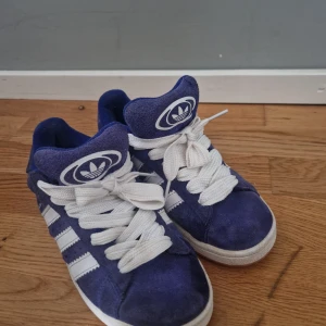 Adidas Campus sneakers blå mocka - Säljer ett par Adidas Campus sneakers i blå mocka med vita detaljer och klassiska tre ränder på sidorna. Skorna har vit sula, breda vita skosnören och Adidas logga på plös och häl. Perfekta för dig som gillar retrostil och streetwear.